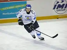 An ice hockey player standing directly in front of the camera. He is wearing a black helmet and uniform.