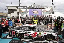 a parked race car covered with confetti and surrounded by celebrating team members