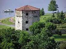 a colour photograph of a stone tower with a red tile roof near a river