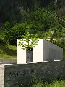 Photograph of a memorial, a simple rectangular structure made of white and grey materials, located at the foot of a green mountainside. In the foreground, part of a stone wall with the inscription 'Gedenkstätte 11.11.2000' is visible.