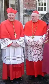 Cardinals Walter Kasper (left) and Godfried Danneels (right) wearing their choir dress: scarlet (red) cassock, white rochet trimmed with lace, scarlet mozetta, scarlet biretta (over the usual scarlet zucchetto), and pectoral cross on a cord.