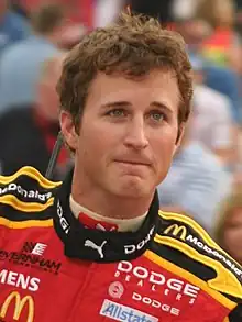 A man in his late twenties wearing black sunglasses with a head full of hair and a red jacket with sponsors' logos.