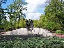 A low stone wall, curving upward. Three statues of Polish soldiers are mounted at its center. Below the statues, Text is mounted as per caption.