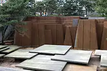 Brown-colored columns standing behind a collection of square-shaped platforms. In the background are trees and other buildings.