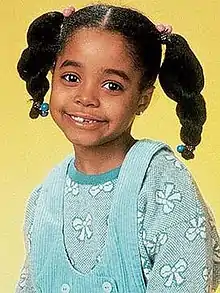 A young African American girl approximately aged five or six smiling at the camera.  Dressed in a blue jumper and overalls, her long black hair is worn in a pair of pig tails that protrude from opposite sides of her head.