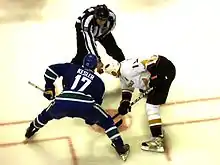 Getzlaf leans in against an opposing player, each with their stick on the ice, as linesman prepares to drop the puck.