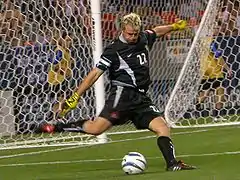 Goalkeeper Kevin Hartman, wearing a black uniform and yellow gloves, prepares to kick a stationary soccer ball near the goal line.