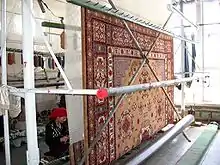 Vertical factory loom seen from the back. Weaver sits down left.