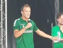 Fair-haired white man in a green shirt speaking into a microphone