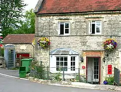 Stone building with old metal sign above the door saying Old Post Office, red post box set into the wall and to the left an old blue telephone box. The door is below the level of the road and separated by metal railings. Hanging baskets of flowers on the front of the building.