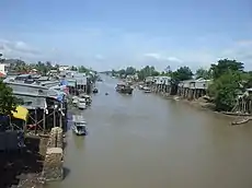 Brown-coloured waterway, with many small boats and elevated wooden and thatched houses visible. Foliage is also in abundance.