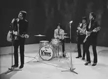 Four members of the band the Kinks stand onstage, during a television appearance. From left to right is Ray Davies, wearing a leather suit and long black pants. He is strumming an acoustic guitar and preparing to sing into a microphone placed in front of him. Next is Mick Avory, seated and playing the drums. He is wearing a bright coloured long sleeve shirt and dark pants. After him is bassist Pete Quaife, playing his instrument and wearing set of clothes similar to Avory's. Last is Dave Davies, wearing what appears to be a leather suit with a short tie, black pants and high-heeled loafers. He plays an elaborate v-shaped electric guitar and is standing behind a microphone.