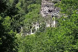 An overgrown, multi-story stone building in ruins. It lies in the mountains, surrounded by forest.