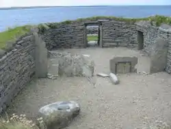 A small area of gravel is enclosed by a stone wall built into the surrounding grassy fields. Various large stones sit inside this enclosure and a low doorway has been constructed at the far end. Beyond the doorway, there is a rocky foreshore and a body of water.