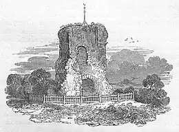 A ruined tower with two arched openings, one at ground level and one immediately above. The tower is surrounded by a low fence, with trees in the background.