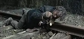 A man with dirty and tousled hair is seen lying on his stomach on a train track, examining a watch that he is holding with his right hand. His shirt is dirty with mud on it and his gloves are torn, baring his fingers. His shoes are also muddy.