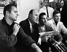 "Four men sit on a couch; three look on as the fourth speaks to a group of reporters off camera"