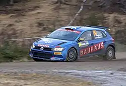  Five-door two-box car in a racing livery fitted with a rear aerofoil and a roof-mounted air scoop for ventilation travelling at speed on a gravel course