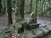 An ancient statue of a young boy riding a cow and a horse at the same time by straddling both at once. The boy is an early emperor on the Kumano pilgrimage. It is set within the forest and is near sacred Buddhist sites.