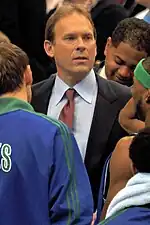 A man, wearing a black suit, blue shirt and red tie, is standing in the middle of a huddle.