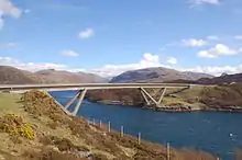 A concrete road bridge spans a body of deep blue water with brown hills beyond