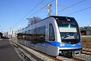 A Blue and gray train with black glass stands idle with overhead wires and adjoining building visible.