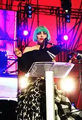 A woman in front of a podium, wearing a one shouldered black top and a black-and-white flowing skirt. She wears a blue bob wig and smiles towards the camera