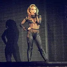 Lady Gaga standing on a stage in a mesh leather cut-out bodysuit and knee-high leather boots. Half of her hair is dyed green.