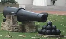 A long 19th-century cannon with a stack of cannonballs in front of it, sitting on a lush green lawn.