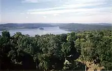 View across forest towards a lake surrounded by low, forested hills. Two small islands are set close to the far shore. There is a small clearing in the immediate foreground. On the left side of the far shore is a wide grassy region set against the forested hills.