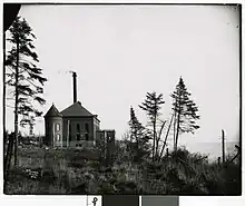 Black-and-white photo of a Romanesque revival building along a lakeshore with pine trees