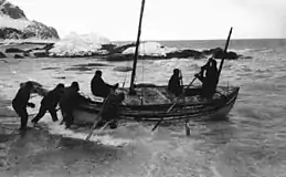  A group of men pushing a boat from a rock beach into the sea, with a background of rocks.