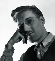 Black-and-white photograph of a man in glasses resting his head on his hand and smiling at the camera