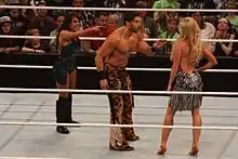 A brunette Caucasian woman and dark-haired Caucasian man in a wrestling ring, pointing and yelling at a blonde Caucasian woman with her back to the camera.