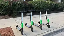 Four electric stand-up scooters lined up on a sidewalk ready for rental. These have black handlebars, white stems, a green battery pack, and gray decks.