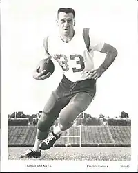 Black-and-white photograph of Infante wearing a football uniform with pads (but no helmet), with a number 33 jersey, and cradling a football in his right arm