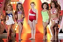 A film screenshot showing five girls standing on stage at the pageant at the end of the film. From left to right, the first girl (wearing a one-piece swimsuit) has her left leg placed in front of her right, the second girl (wearing a two-piece swimsuit) has her legs shifted in the opposite direction, at center the main character (wearing a one-piece swimsuit) is standing straight, the fourth girl (wearing a one-piece swimsuit) has her left leg placed in front of her right, and the last girl (wearing a two-piece swimsuit) also has her left foot forward. All of the girls are smiling towards different angles. The background of the stage is decorated and it is reflected on the shiny surface of the stage.