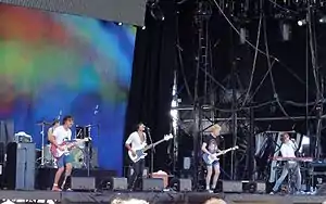 A distant shot of five men performing on a stage. The man at extreme left is behind a drum kit and is mostly obscured by the next man. He is singing and playing a guitar. The third man is also singing and plays a bass guitar. The fourth man plays a guitar and sings. The fifth man is behind a keyboard and holds the microphone as he sings.