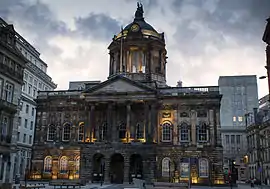 A neoclassical building in two storeys with a central portico and a dome seen almost end-on.  It is flanked on each side by other buildings