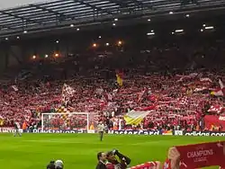 A stand which is full of people standing to support their team. There are a number of flags, scarfs, and banners in the crowd.