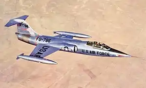 Jet fighter in metallic scheme with T-tail and short wings flying above desert and black constructions