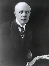 A balding, white-haired man wearing 1920s formal business attire (jacket and waistcoat with wing collar and tie) leans to his left on a table and looks directly at the viewer. His right hand rests in front of his left on the edge of the table next to some papers and a pen.