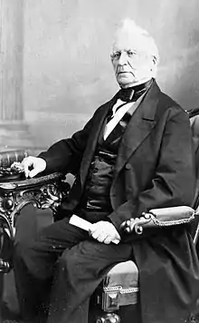 Photo of fair-skinned man with white hair, tuft in front, wearing mid-Victorian business suit, sitting at a table