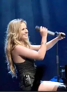 Lucie Silvas wearing a black sleeveless top and black shorts, sitting behind a piano onstage, grabbing a microphone from a stand, and grinning