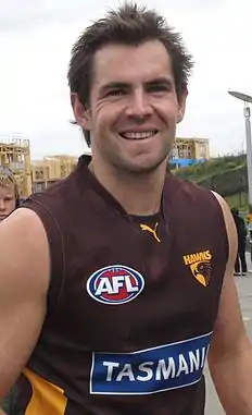 A male athlete with dark hair wearing a sleeved jersey smiles at camera.