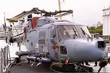 A light blue helicopter on a ship's flight deck with its rotor blades folded away. A large dark blue missile is attached to its right side and the number 335 is written in white on its door.