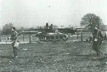 A Locust tank, facing right, on a road that cuts through a field. Several uniformed men move in the opposite direction in the foreground. A large military glider can be seen in the background, just behind the Locust.