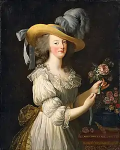 Three-quarter length portrait of a woman wearing a large straw hat with grayish-blue ostrich feathers and a ruffly cream dress with a gold bow tied at the waist in the back. She is arranging a group of pink flowers.