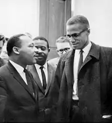 Malcolm X and Martin Luther King Jr. speak to each other thoughtfully as others look on.