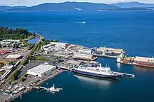 One large ship and one small ship are docked at a port. There is a large expanse of water with another small ship approaching and mountains in the background.